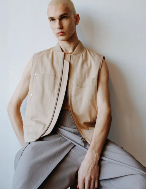 Portrait of an adult male model wearing a tan vest and grey trousers, sitting indoors against a plain white wall in soft light.
