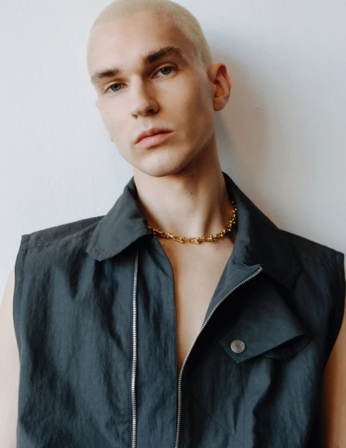 Close-up shot of a blond male model in a sleeveless black shirt and a gold necklace posing against a white wall indoors.
