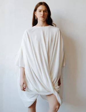 Model Mica Tosi wearing an oversized draped off-white dress and gold drop earrings against a neutral studio backdrop.