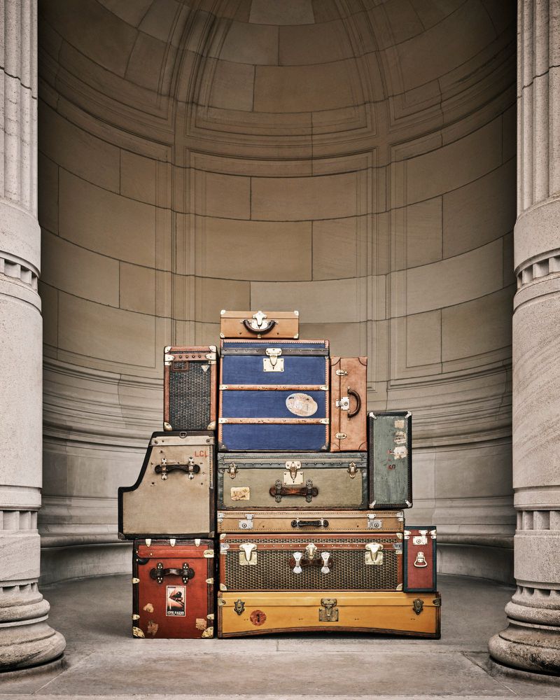 Stack of vintage suitcases and trunks in front of a monumental stone wall and pillars, creating a sense of travel and nostalgia.