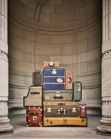 Stack of vintage suitcases and trunks in front of a monumental stone wall and pillars, creating a sense of travel and nostalgia.