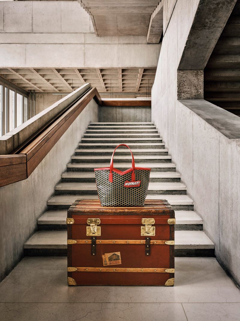 Brown vintage trunk topped with a modern patterned tote bag, set against a concrete stairwell in a minimalist interior space.