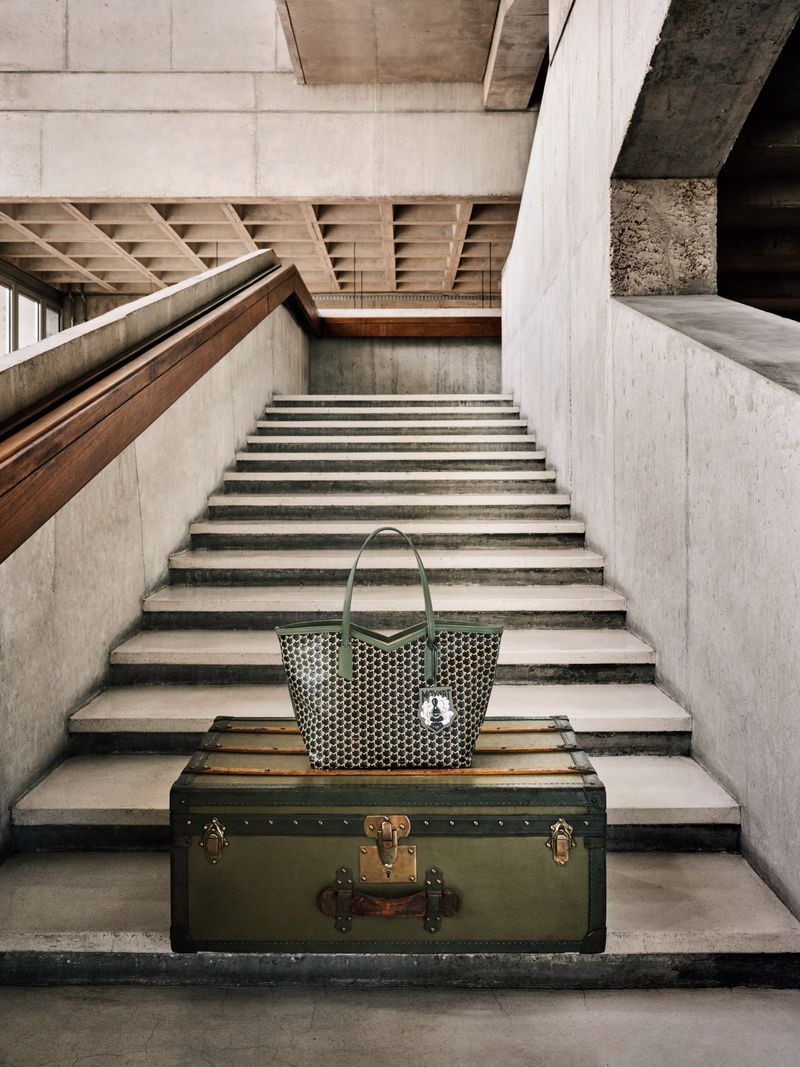 Olive green tote bag on a vintage trunk near a concrete staircase. Interior shot with neutral tones and strong geometrical shapes.