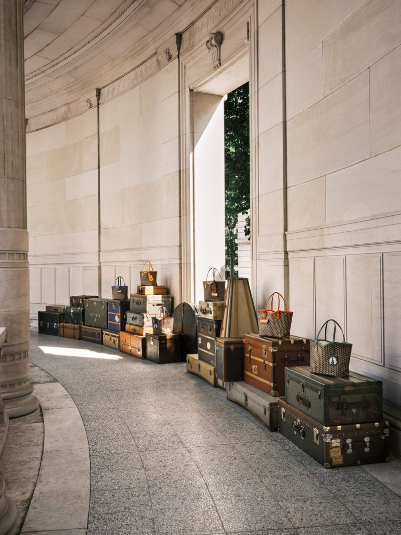 Collection of vintage suitcases and bags stacked in an arched stone corridor, bathed in natural light, creating a travel-themed display.