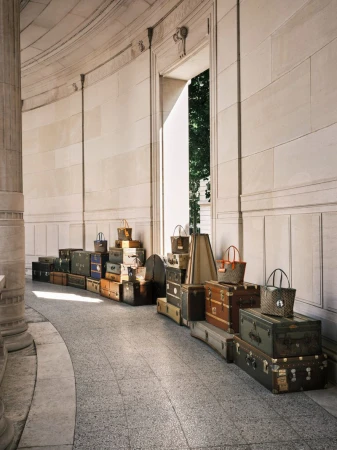 Collection of vintage suitcases and bags stacked in an arched stone corridor, bathed in natural light, creating a travel-themed display.