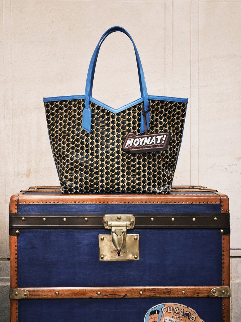 Luxury black patterned tote bag with blue trim sitting on top of a blue vintage steamer trunk in front of a neutral wall.