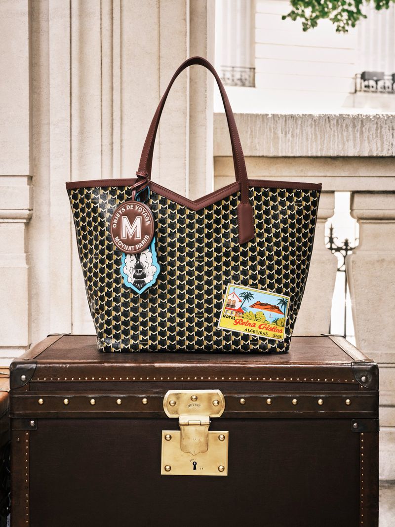 Patterned tote bag with brown leather handles and patches on a dark brown trunk against a light stone architectural background.