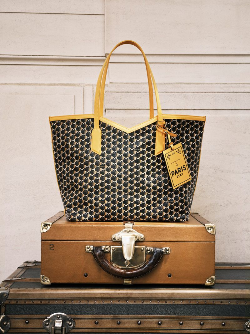 Patterned handbag with yellow handles, top trim and tag on a brown suitcase with silver hardware against a light-colored wall.
