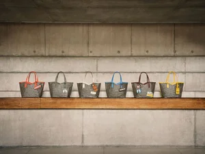 Six patterned Moynat tote bags with multicolored leather handles displayed in a row on a wooden shelf against a concrete wall.