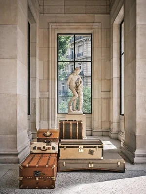 Vintage Moynat leather trunks and luggage stacked on a stone floor in front of a tall window and classical statue.