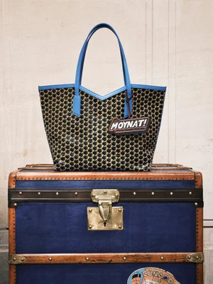 Moynat monogram tote bag with bright blue leather straps displayed on top of an antique blue steamer trunk.