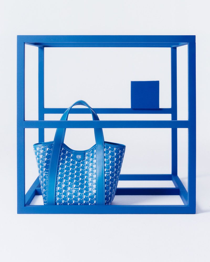 Blue patterned tote bag on a blue modernist display stand against a white backdrop in a fashion or product photography studio.