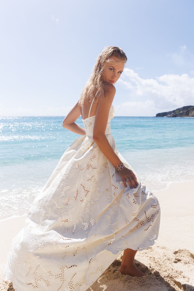 Woman in white dress posing on beach with ocean backdrop. Fashion shoot featuring female model, blue sea, light sky, and sand.