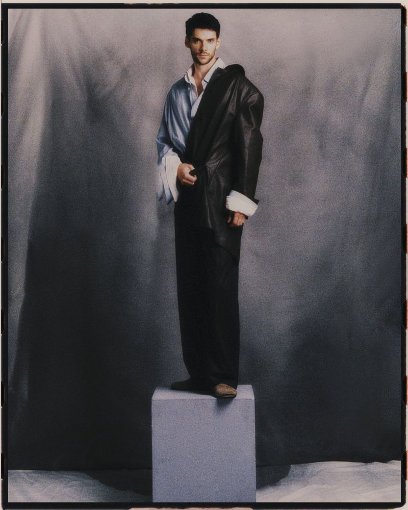 A man in an unbuttoned blue shirt and dark pants stands on a gray cube. He has a black coat over his shoulder in a studio setting.