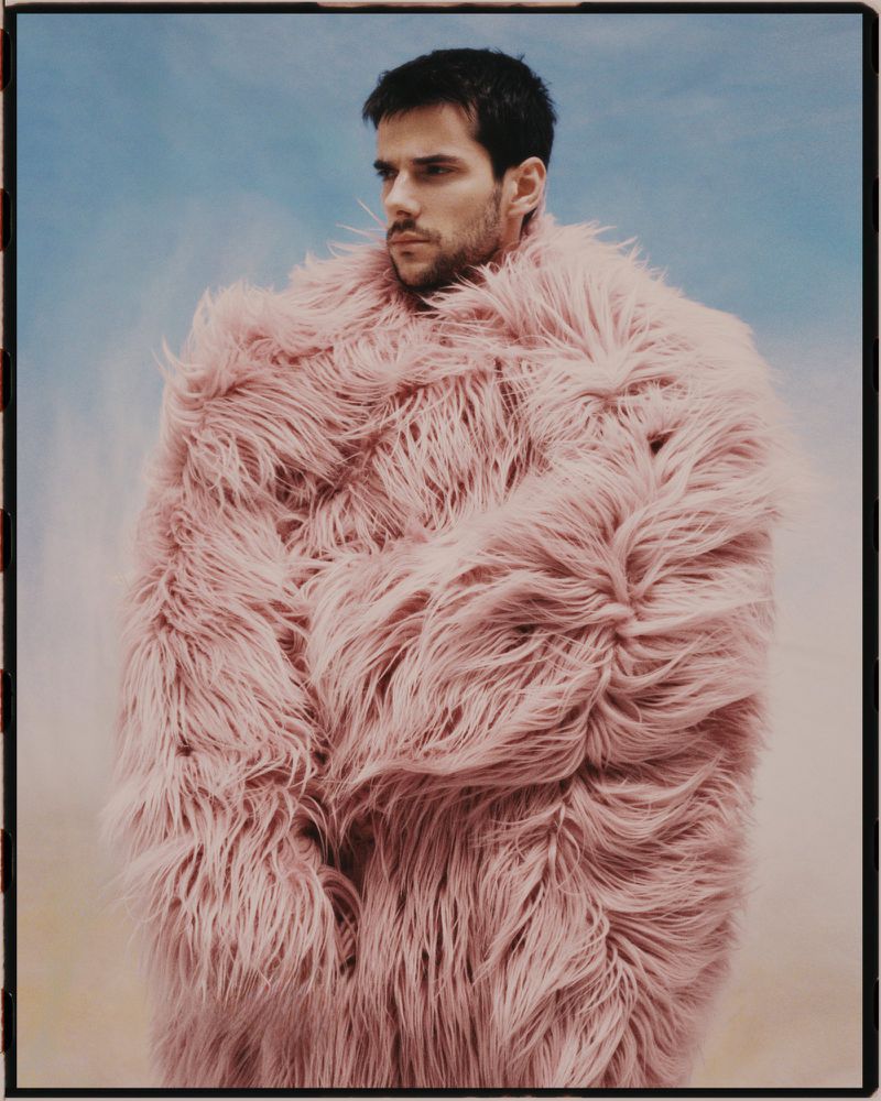 Portrait of a man with dark hair and beard, wrapped in a large, fluffy, light pink faux fur coat against a soft blue background.