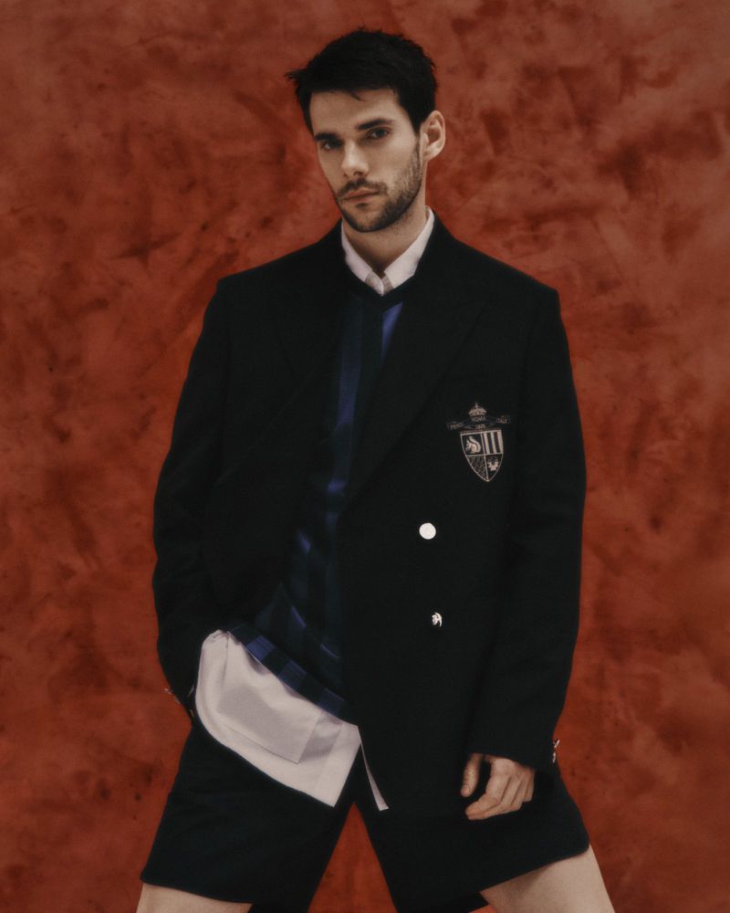 Man in a black blazer, white button-down, and black shorts posing against a textured orange backdrop in a fashion editorial shot.