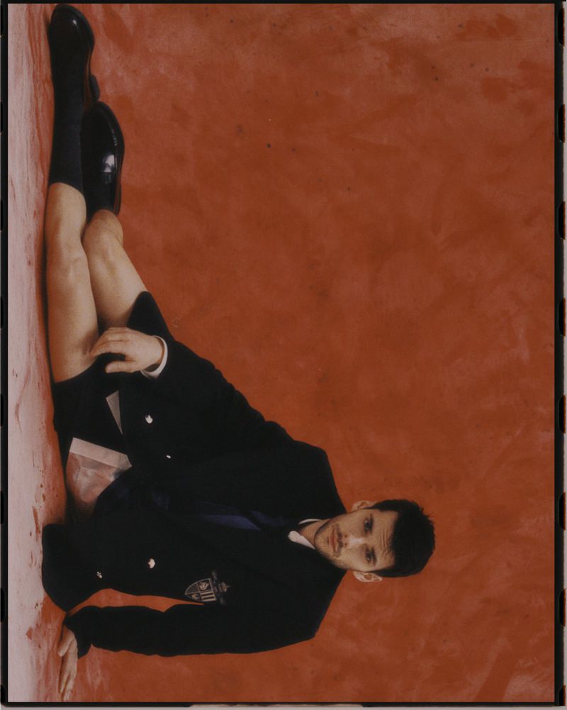 Adult male in a black suit poses against an orange-red backdrop, lying on his side with legs raised. He wears dark dress shoes and black socks.