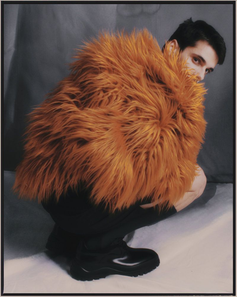 A man with dark hair and complexion crouches indoors wearing a large, orange faux fur coat and black boots, looking directly at the camera.