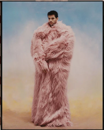 Adult male poses in a full-body pink faux fur coat against a soft blue and gold backdrop in a fashion or editorial style photograph.