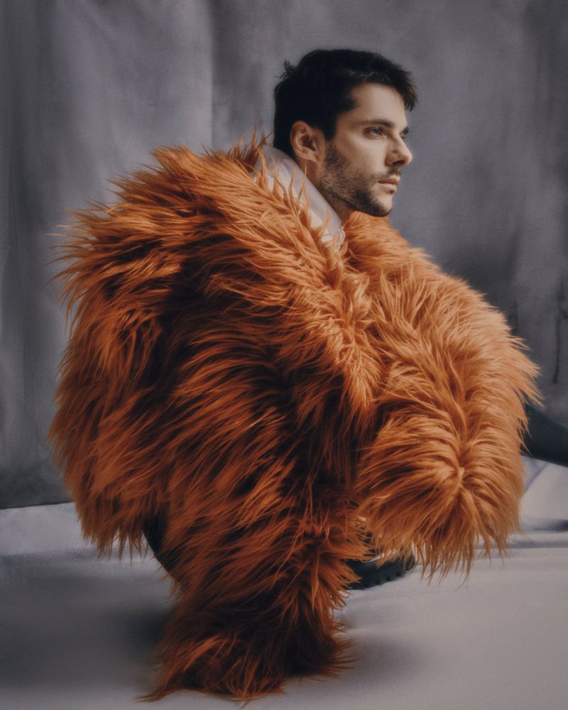 Profile portrait of an adult man wearing a textured, shaggy orange coat, facing right, against a mottled grey studio background.