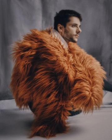 Profile portrait of an adult man wearing a textured, shaggy orange coat, facing right, against a mottled grey studio background.