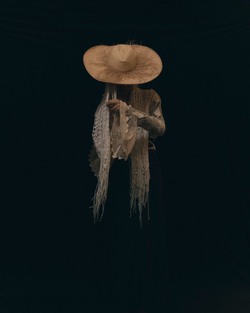 Person in a flowing white lace shawl and wide brimmed hat stands against a dark background. Fashion design with texture and pattern.