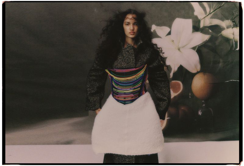 A mixed-race woman with curly hair poses in a textured coat with colorful stripes, a white skirt, against a floral background.
