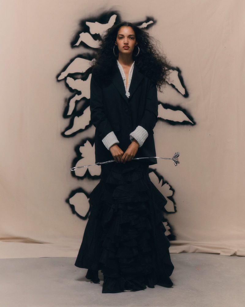 Elegant woman in a black jacket and layered skirt poses against a backdrop with a floral motif, holding a silver ornamental stem.