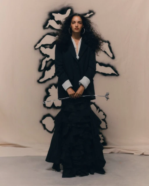 Elegant woman in a black jacket and layered skirt poses against a backdrop with a floral motif, holding a silver ornamental stem.