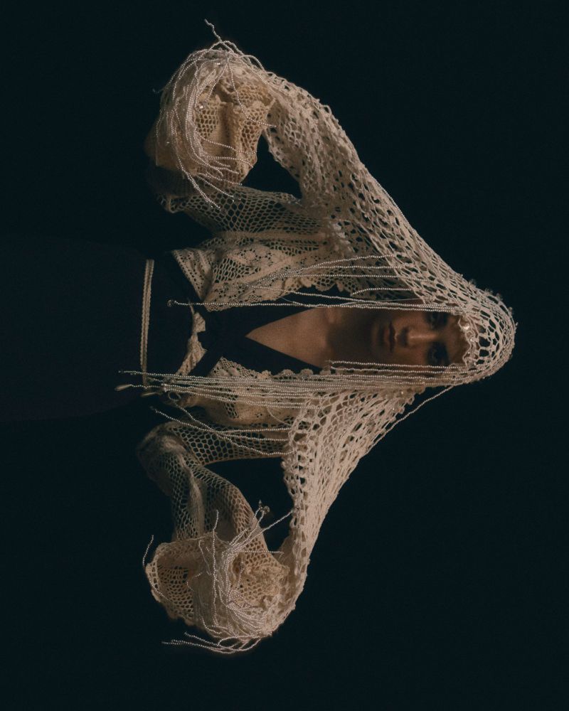 Close-up portrait of adult female under a cream-colored crocheted scarf, against a dark background, enhancing contrast and detail.