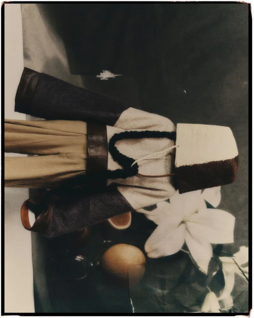 Still life featuring neutral-toned fabrics, a leather sleeve, an orange, and a white flower on a reflective surface, captured in soft lighting.