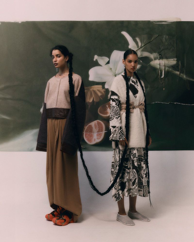 Two young women in ethnic fashion stand in front of a floral backdrop, each adorned with a long braid extending onto the ground.