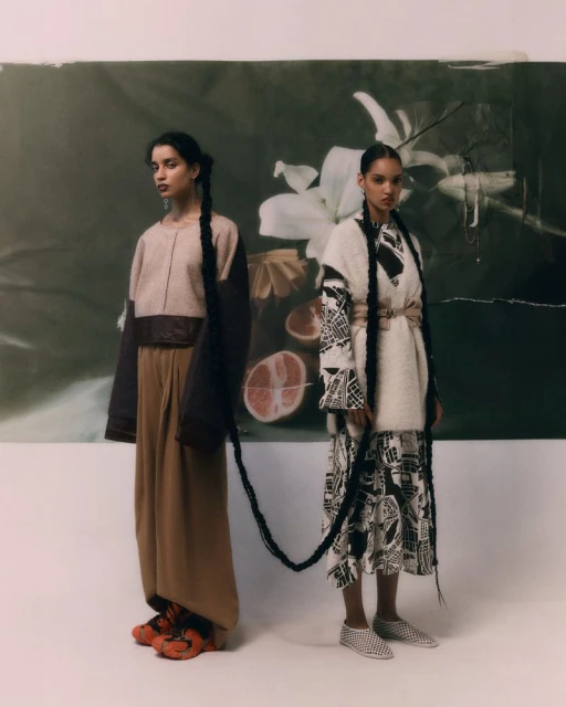 Two young women in ethnic fashion stand in front of a floral backdrop, each adorned with a long braid extending onto the ground.