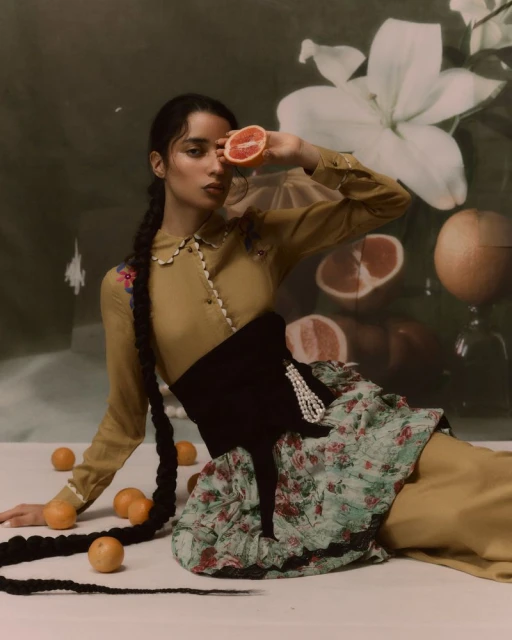 An elegant woman with a long braid and a floral skirt poses indoors with oranges, holding a grapefruit half in front of her face.