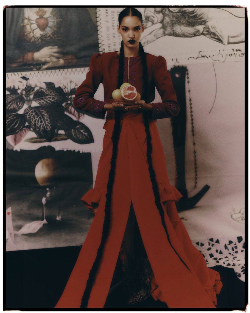 Fashion shot of a woman in a red dress holding citrus fruit in a photo studio against a backdrop of layered art and drawings.