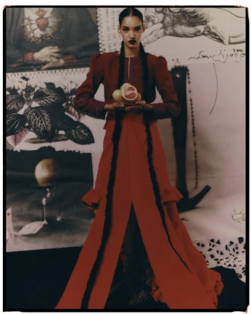 Fashion shot of a woman in a red dress holding citrus fruit in a photo studio against a backdrop of layered art and drawings.