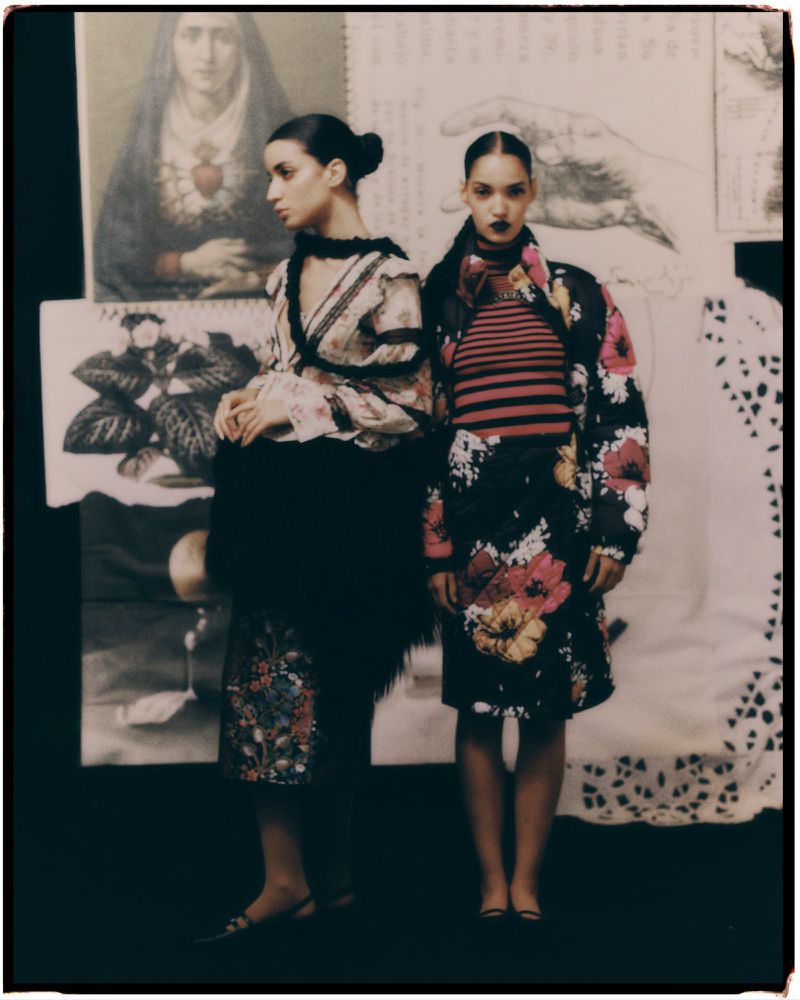 Two models pose in floral print fashion in front of patterned backdrops. One model wears a pink striped shirt and floral skirt.