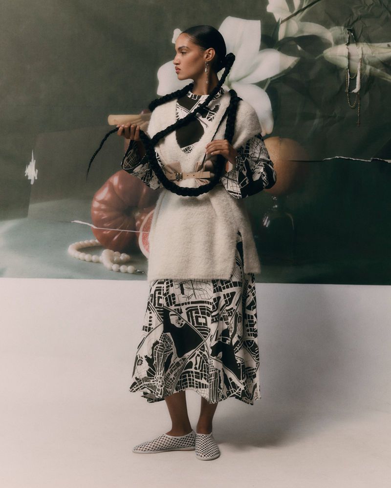 A young adult female model poses in a studio wearing a patterned dress and vest, with a painterly backdrop and soft lighting.