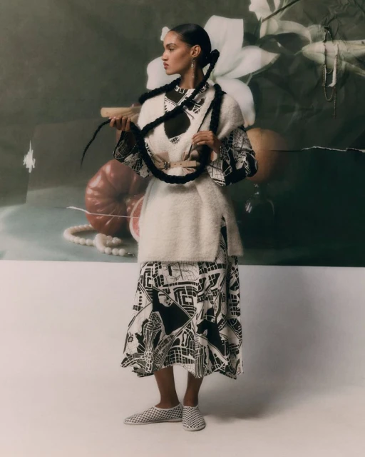 A young adult female model poses in a studio wearing a patterned dress and vest, with a painterly backdrop and soft lighting.