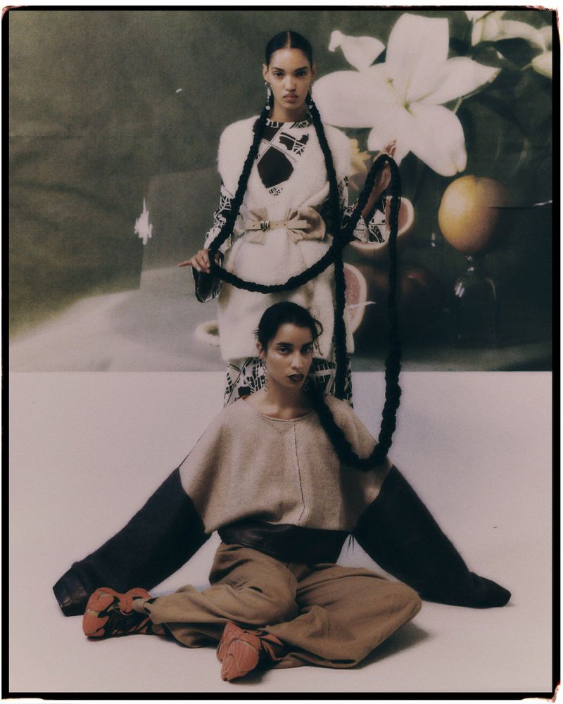 Two women pose in a fashion editorial shot, showcasing beige and brown clothing, elaborate braided hairstyles, with a painted backdrop.