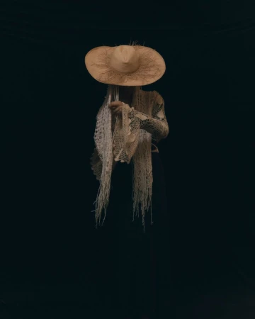 Model Corinne Murray wearing a wide-brim straw hat and cream fringed crochet shawl against a black background.