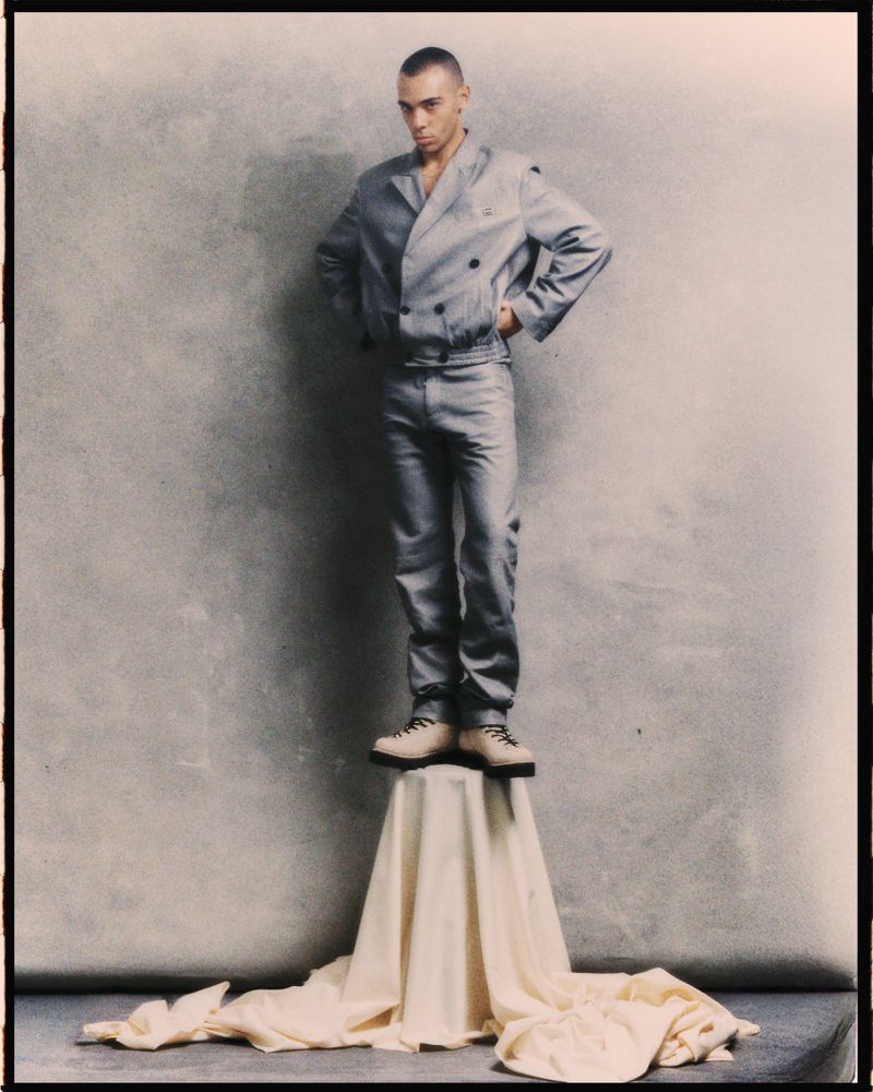 Full shot of a man in a gray suit standing on a draped pedestal against a mottled gray background in a studio setting, fashion shot.