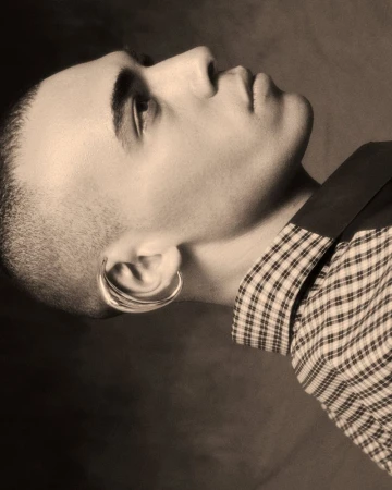 Portrait of a young adult male in profile wearing a plaid shirt and hoop earrings, photographed indoors in a sepia tone.