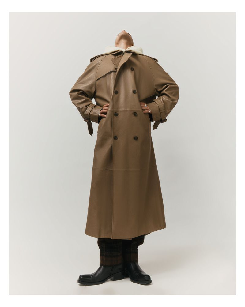 Man in tan leather trench coat standing in a photo studio looking up with his hands on his waist in a fashion editorial.