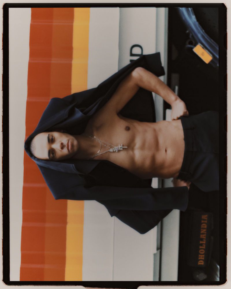 Shirtless male model wearing dark trousers with a dark jacket draped over his head in front of a striped truck exterior.