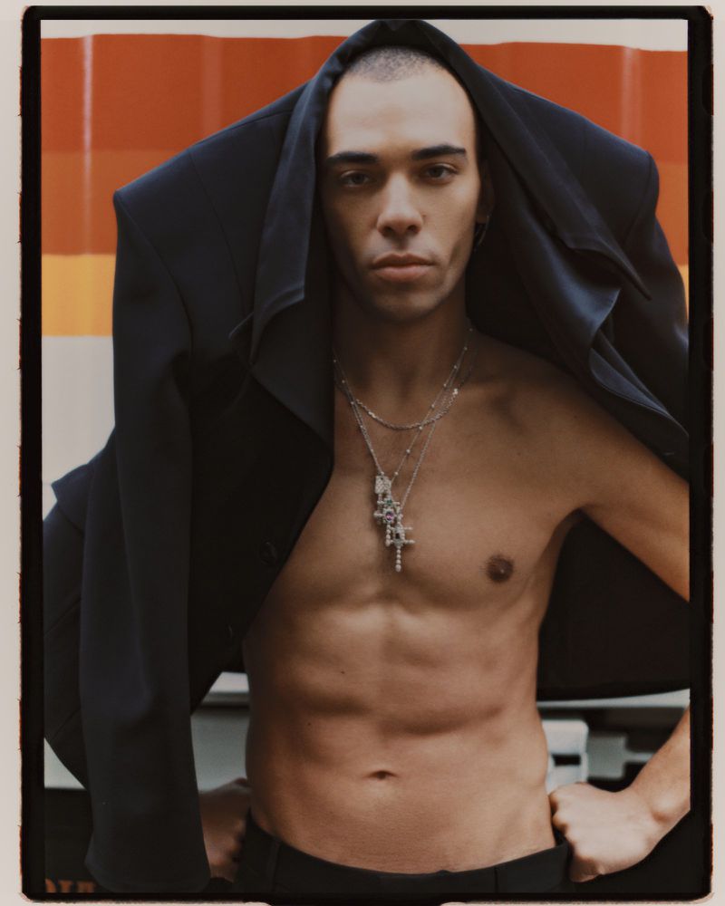Portrait of a shirtless adult man with a blazer draped over his head and silver necklaces standing against a multicolored background.