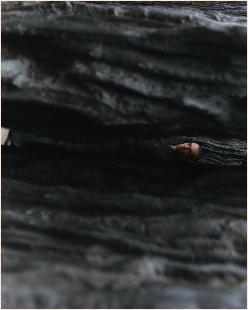 Adult laying between rock formations, possibly in a cave. Person is wearing all black clothing and the rocks are dark gray and layered.