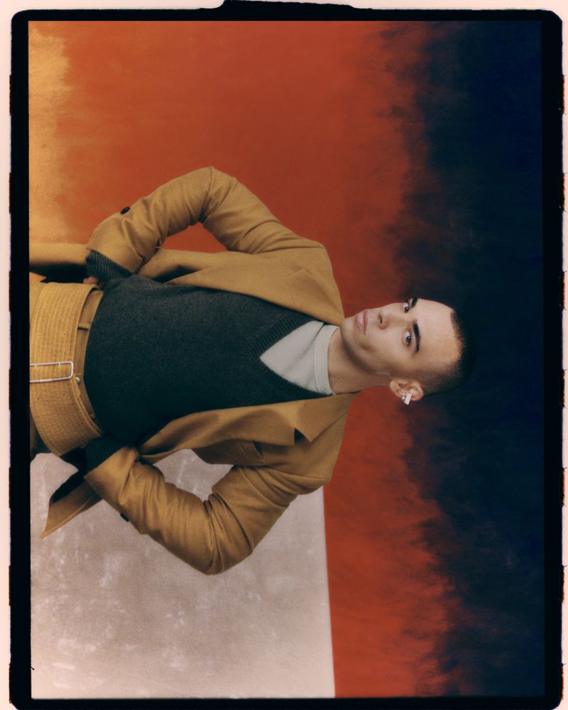 Adult male poses in a tan jacket and pants, a gray sweater, and white earbuds against a warm-toned and gray gradient backdrop.