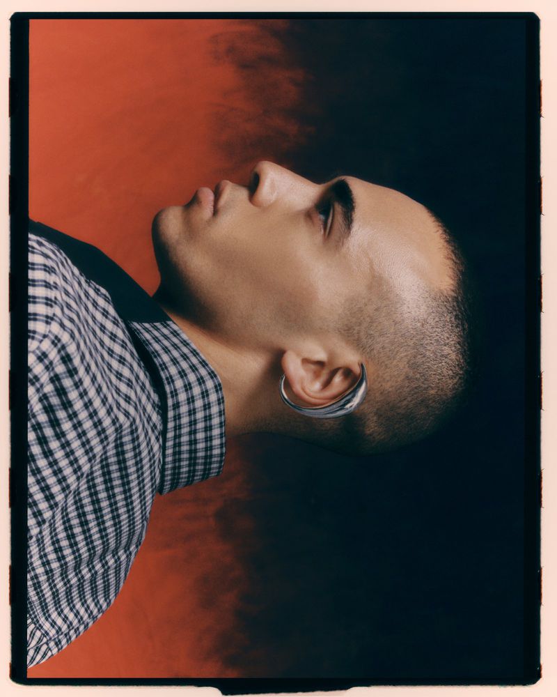 Portrait of a young adult male with short hair and an earring, wearing a checkered shirt, set against a backdrop of diffused orange and black.