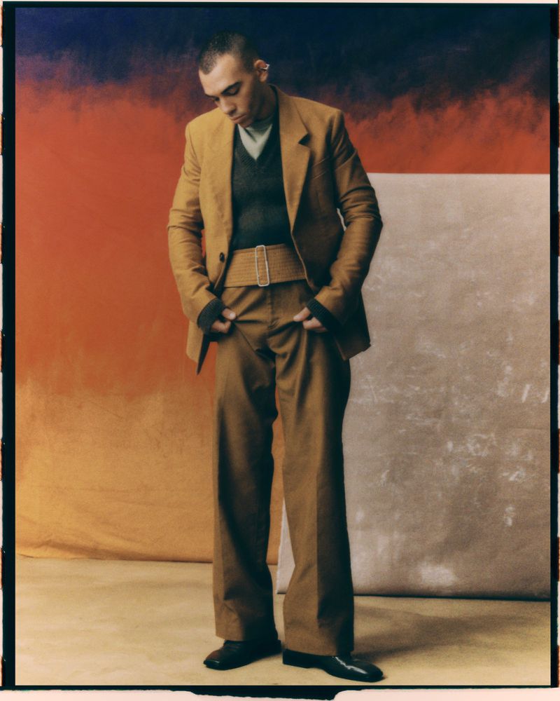 Full-length studio shot of a man in a tan suit with a dark sweater, posing against a backdrop with split orange and white sections.